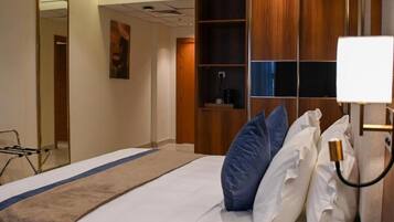 A hotel room with a bed, a bedside table with a lamp, a wardrobe, a mirror, and a wall-mounted shelf.