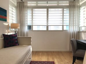 A bedroom with a bed, a desk, a chair, a window with blinds, and a lamp.
