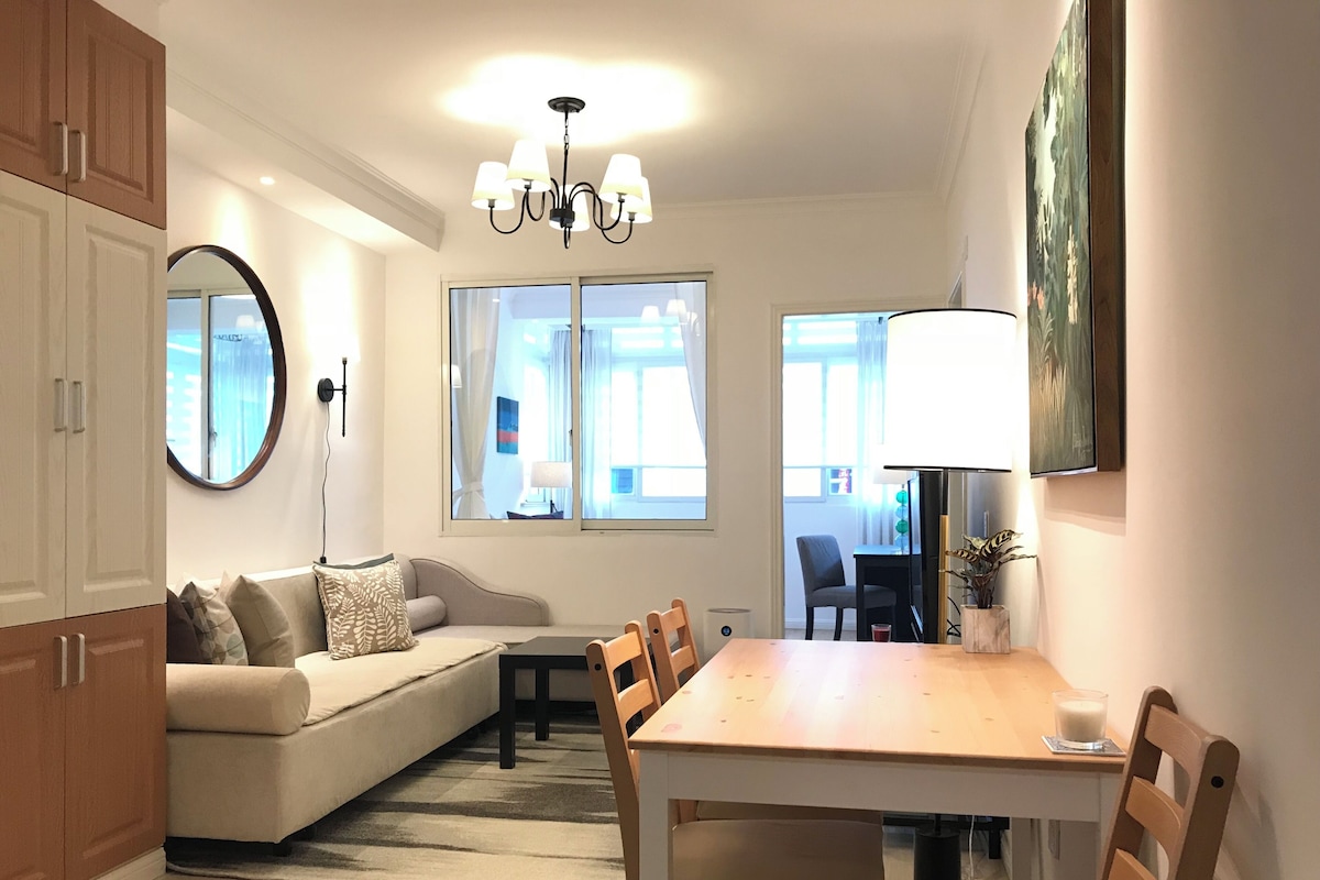 A modern living room with a sofa, dining table, chairs, a chandelier, and large windows.