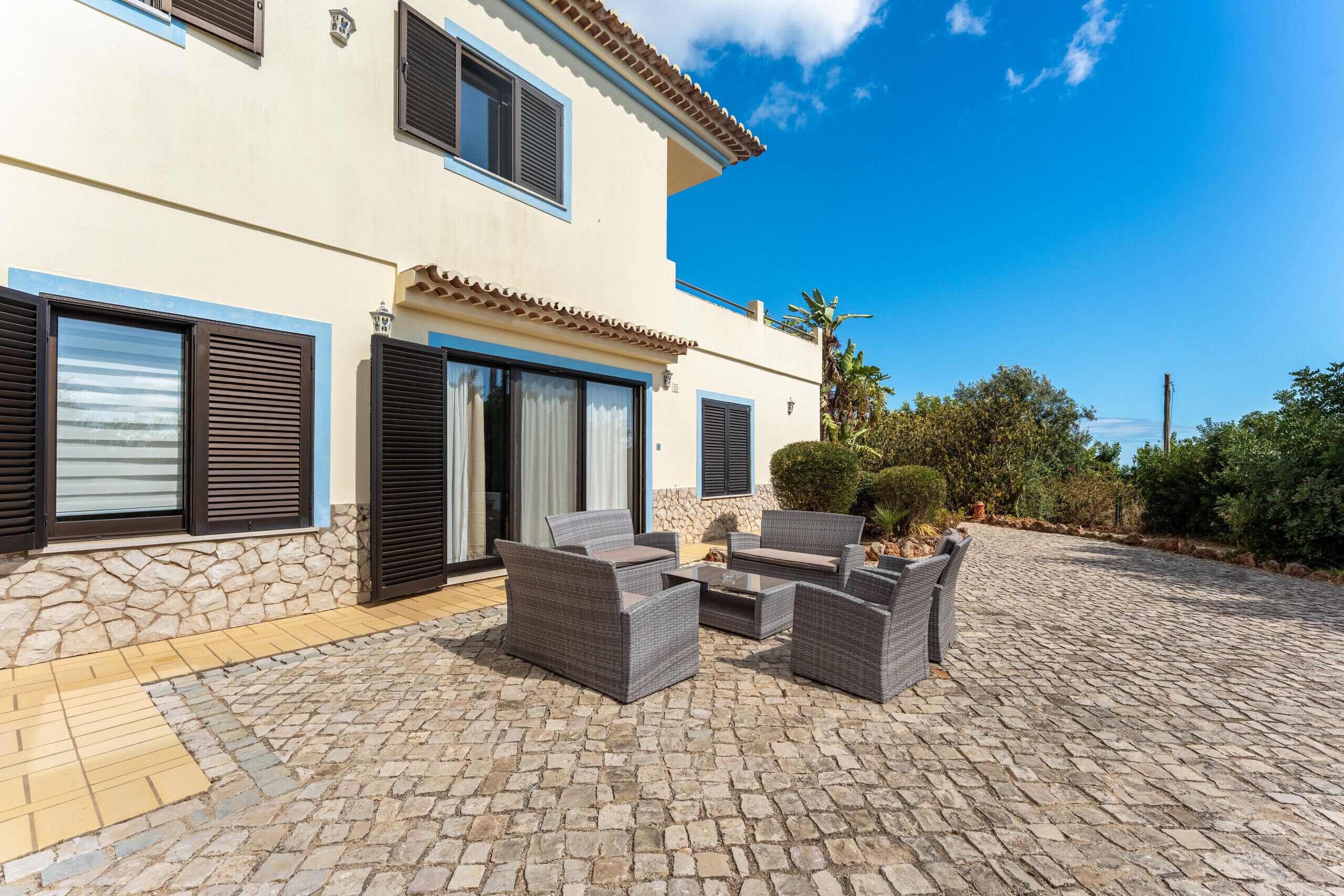 Un edificio de dos pisos con fachada de piedra, varias ventanas con contraventanas y un patio empedrado con mobiliario exterior.
