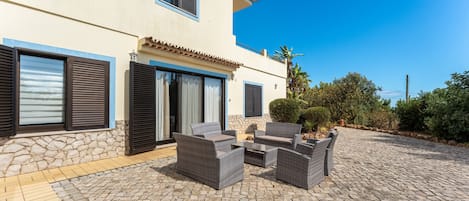 Un edificio de dos pisos con fachada de piedra, varias ventanas con contraventanas y un patio empedrado con mobiliario exterior.