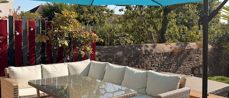 Et udendørs opholdsområde med en fletsofa, et bord med glasplade og en blå parasol.