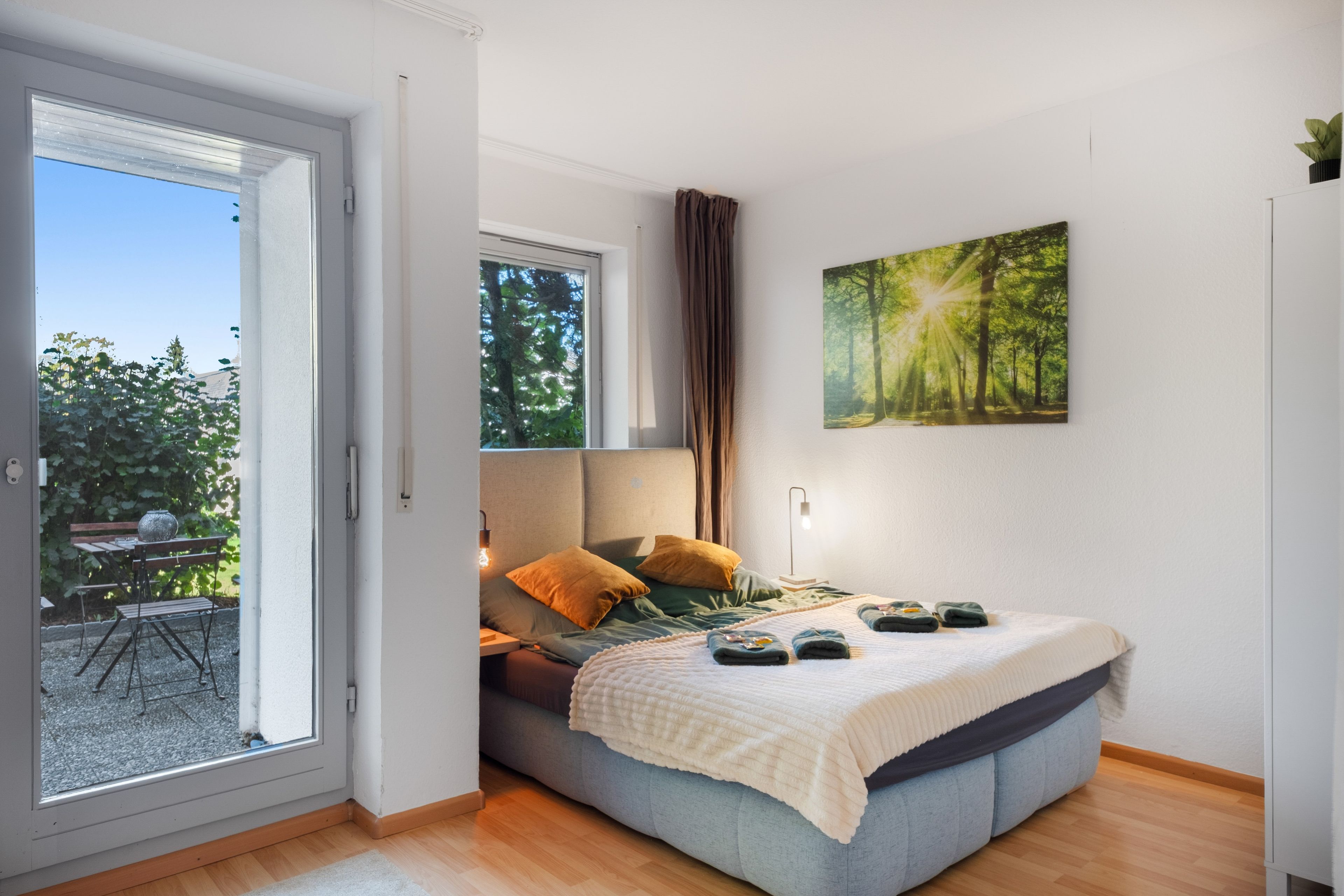 A bedroom with a bed, a window, a painting, and a view of the outdoors.