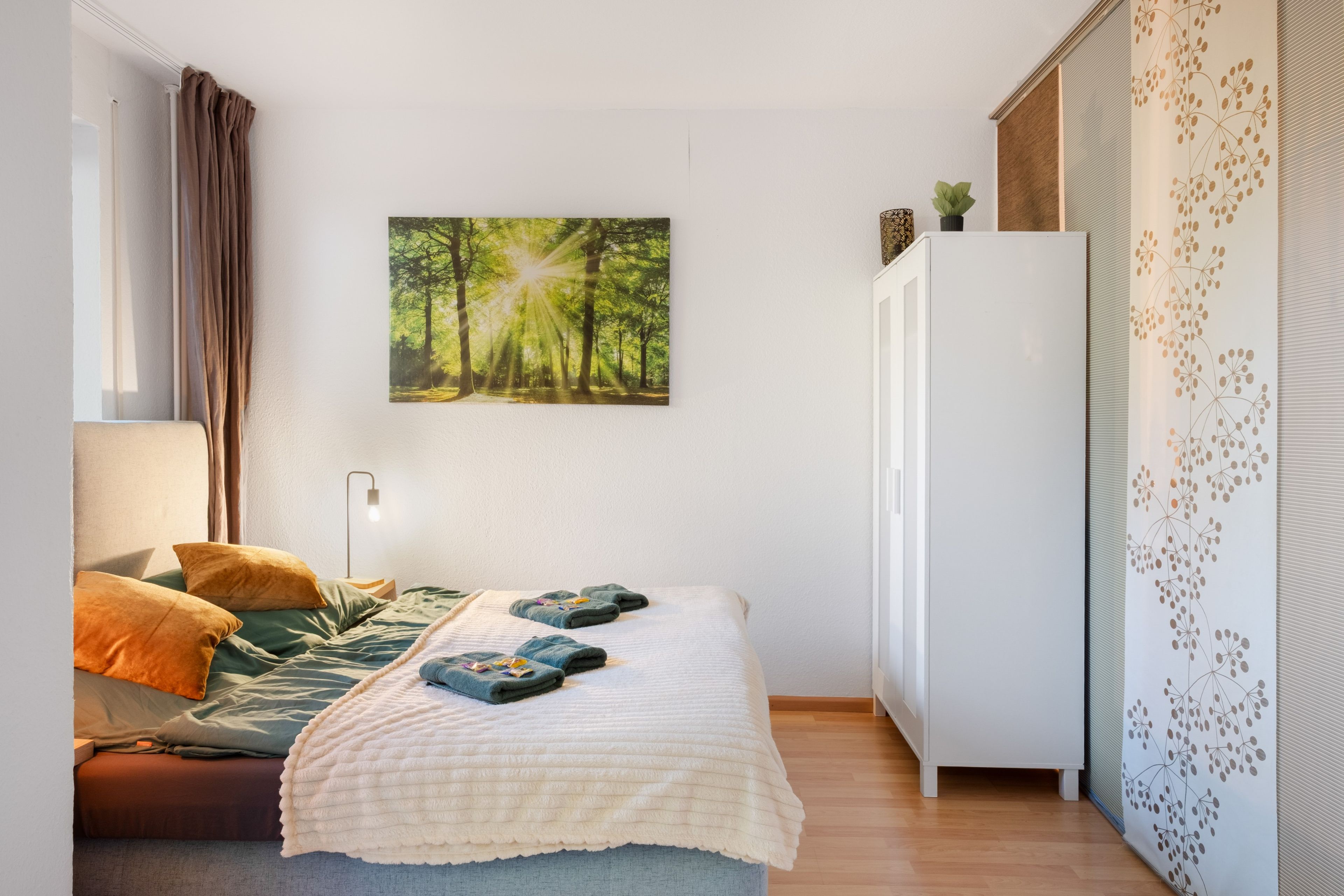 Ein Schlafzimmer mit einem Bett, Kissen, einer Nachttischlampe, einem Kleiderschrank und einem Wandbild von einem Wald.