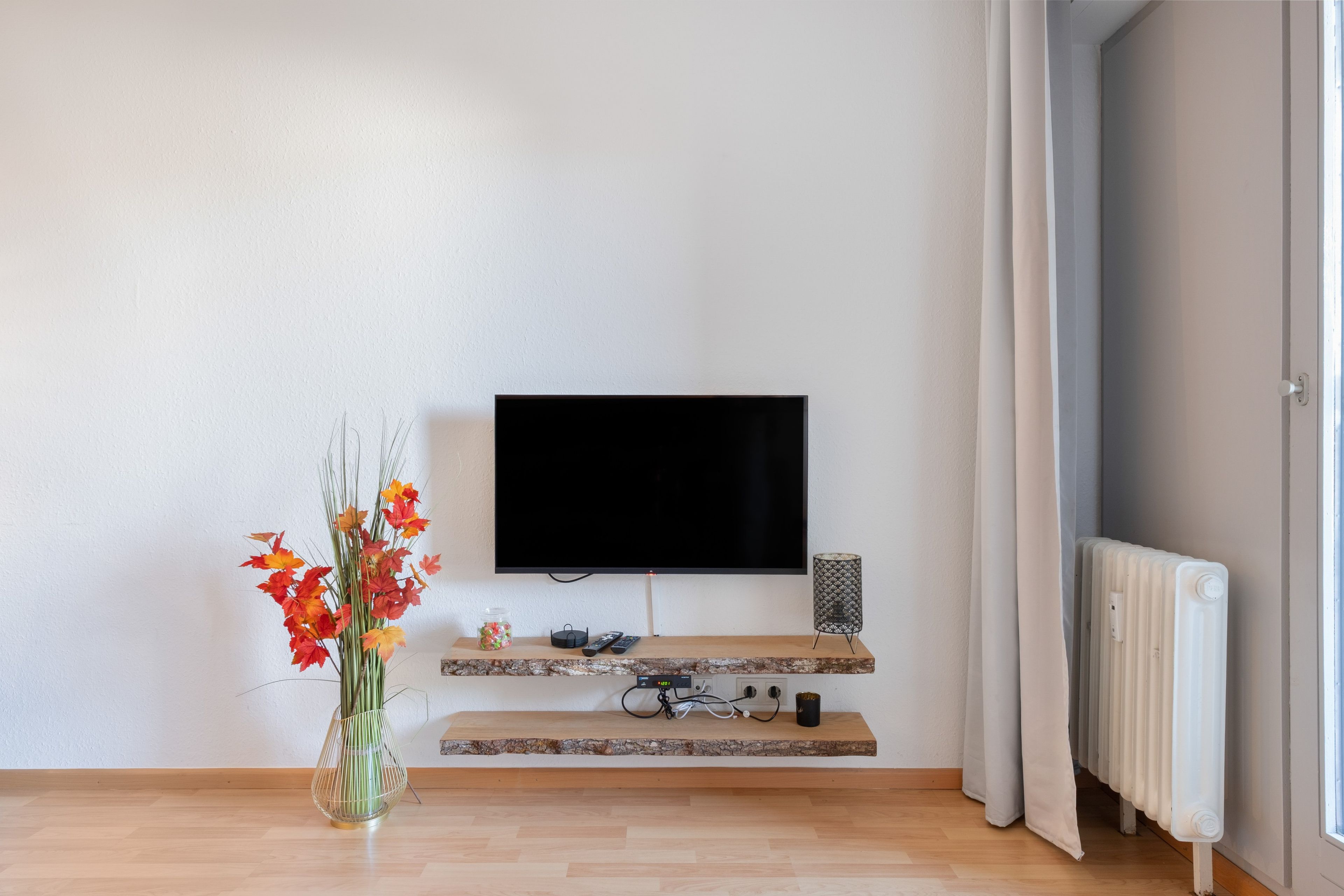 Ein modernes Wohnzimmer mit einem an der Wand befestigten Flachbildfernseher, einem Holztisch, einer Blumenvase und einer Heizung.