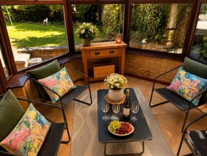 Eine sonnendurchflutete Terrasse mit Esstisch, umgeben von Stühlen mit bunten Kissen, einer Blumenvase und einer Weinflasche.