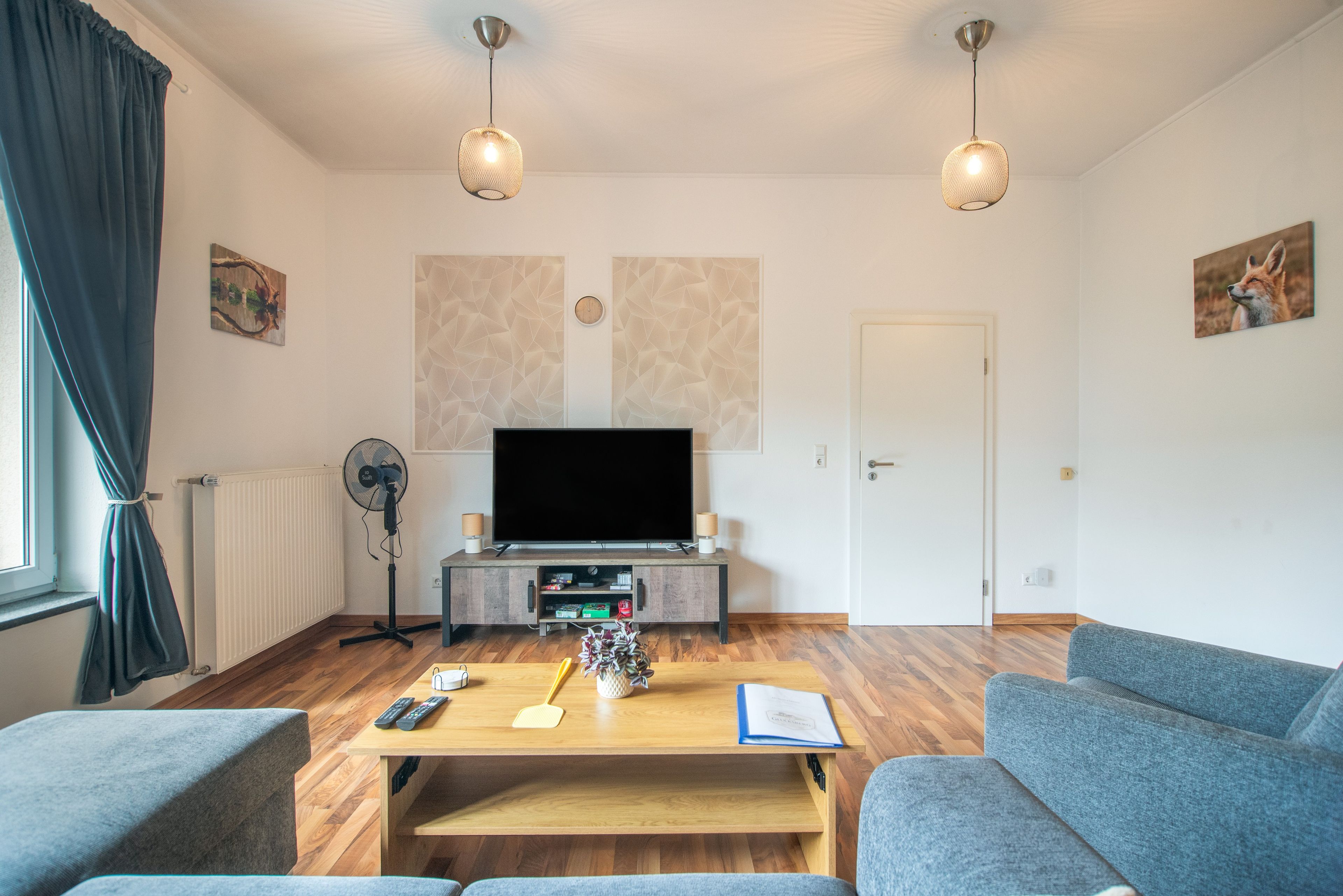Un salon moderne avec un téléviseur à écran plat, une table basse en bois, un canapé gris et deux lampes suspendues.
