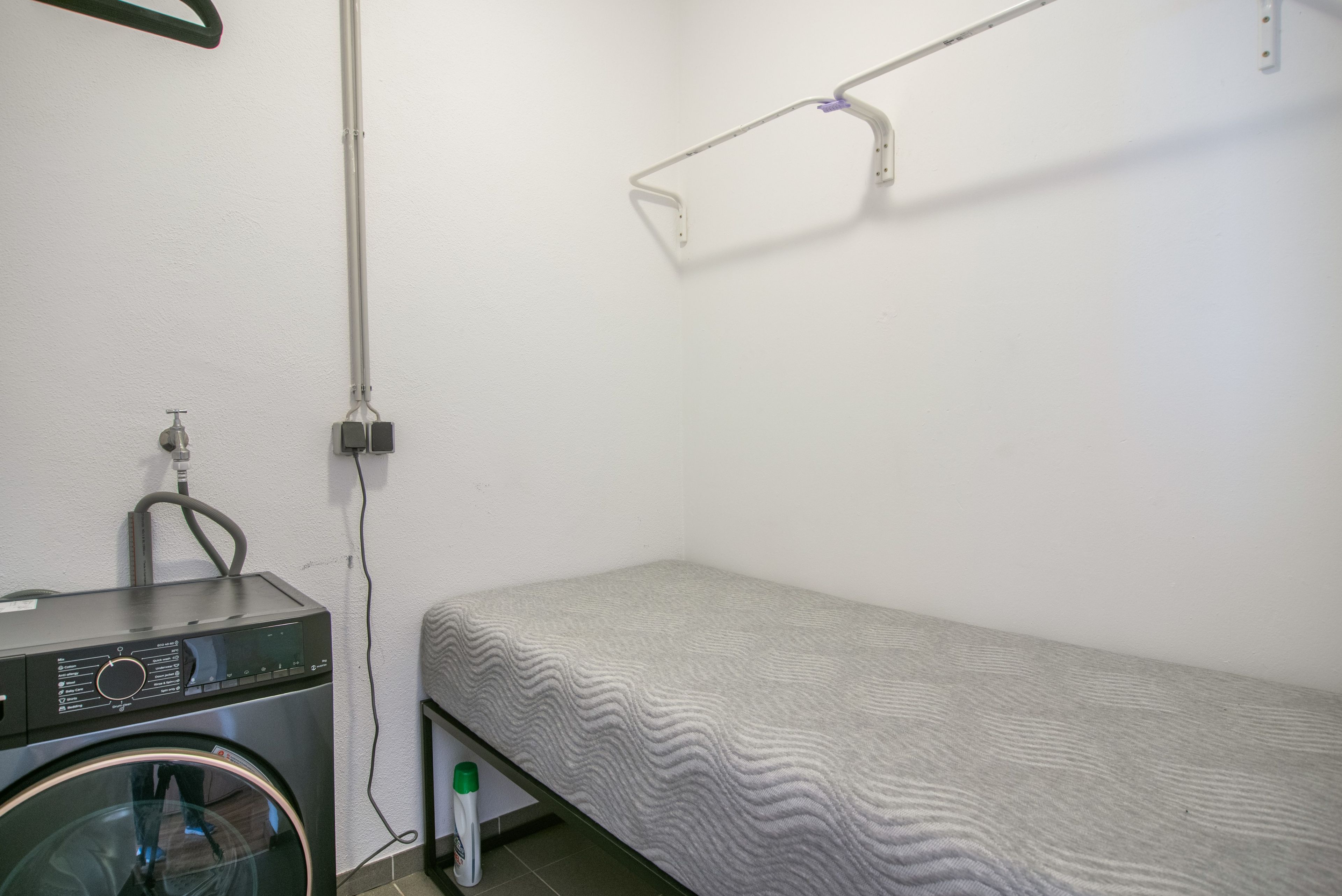 Une petite chambre avec un lit simple, une machine à laver et un étendoir.