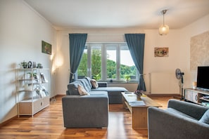 A modern living room with a sofa, armchairs, a coffee table, and a TV. There are windows with curtains, a bookshelf, and a radiator.