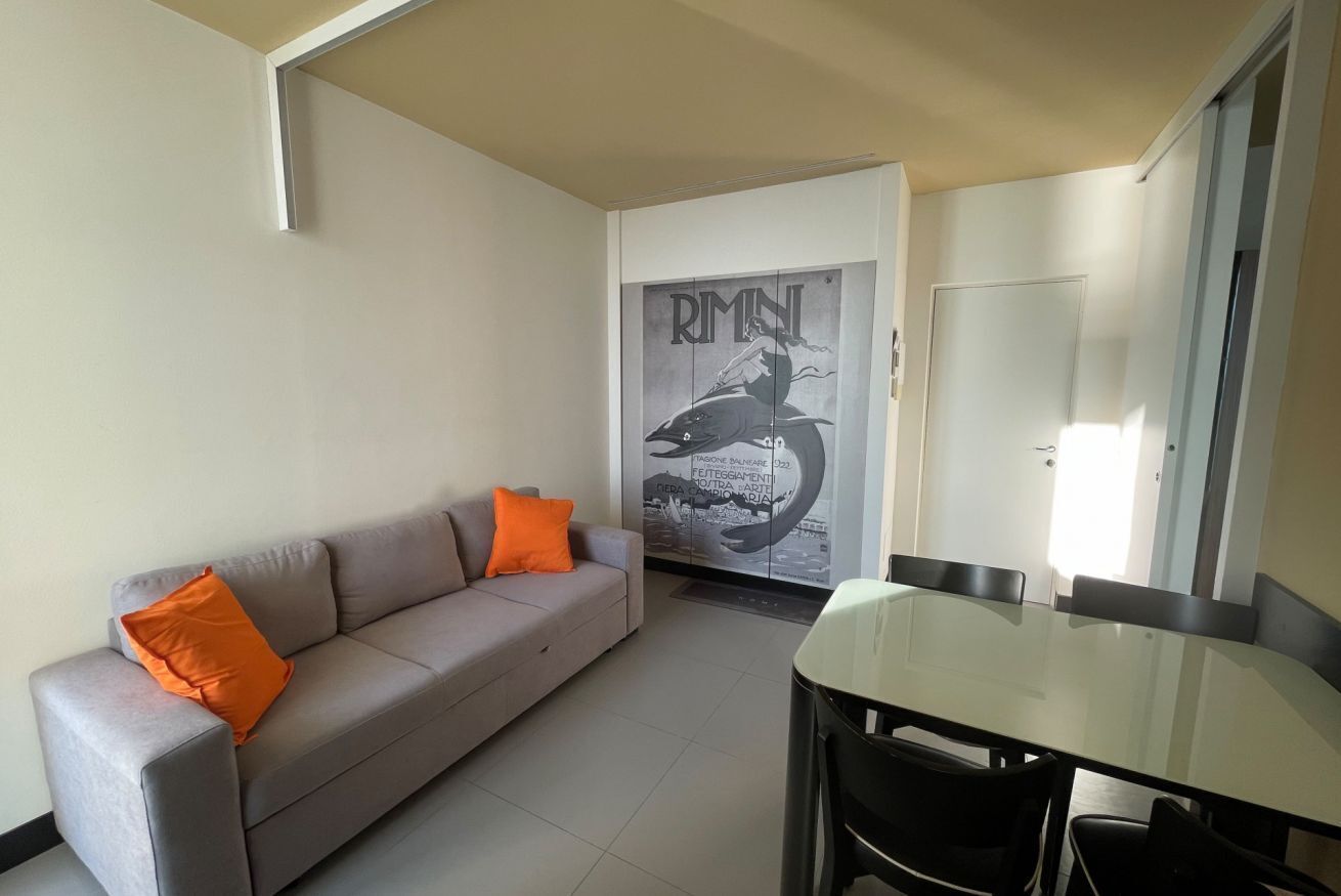 Un salon avec un canapé gris, des coussins orange, une table à manger en verre et une porte ornée d’une affiche.