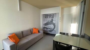 Un salon avec un canapé gris, des coussins orange, une table à manger en verre et une porte ornée d’une affiche.