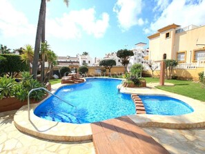 A swimming pool with a wooden deck and a stone border, surrounded by greenery and buildings. - Cozy 3-bedroom house in Villamartin, Orihuela Costa (Orihuela Costa, Alicante)