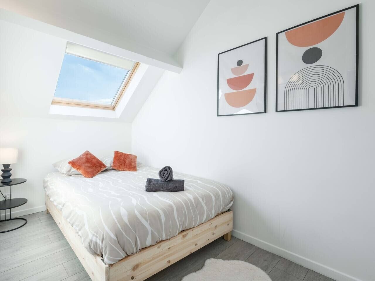 Ein Schlafzimmer mit Dachfenster, einem Bett mit weiß-grauem Muster, zwei orangefarbenen Kissen, einem kleinen runden Tisch mit Lampe und zwei gerahmten abstrakten Kunstwerken an der Wand.