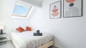 Ein Schlafzimmer mit Dachfenster, einem Bett mit weiß-grauem Muster, zwei orangefarbenen Kissen, einem kleinen runden Tisch mit Lampe und zwei gerahmten abstrakten Kunstwerken an der Wand.