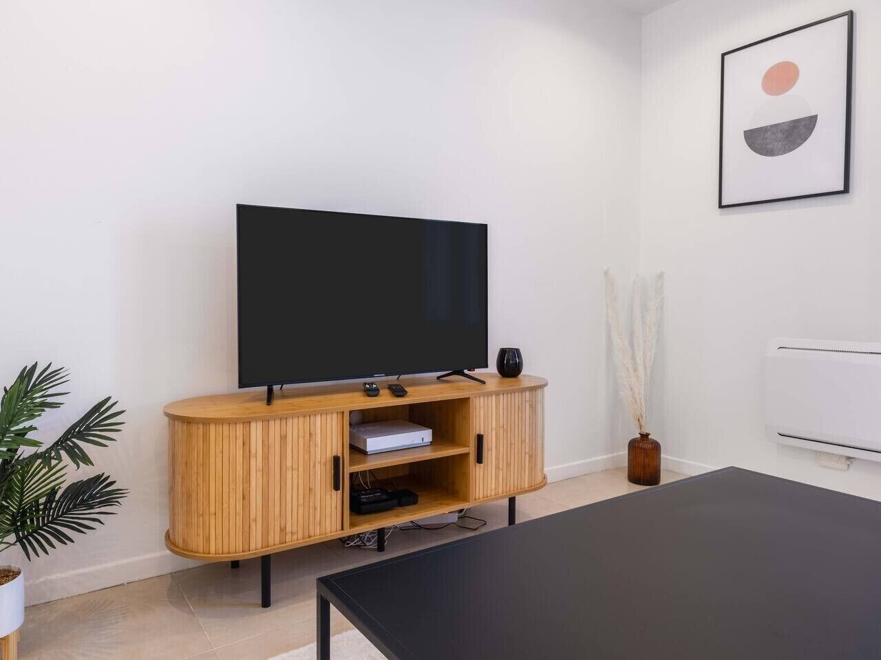 Ein modernes Wohnzimmer mit Flachbildfernseher auf einer Holzkassette, einem schwarzen Couchtisch und einer Topfpflanze.