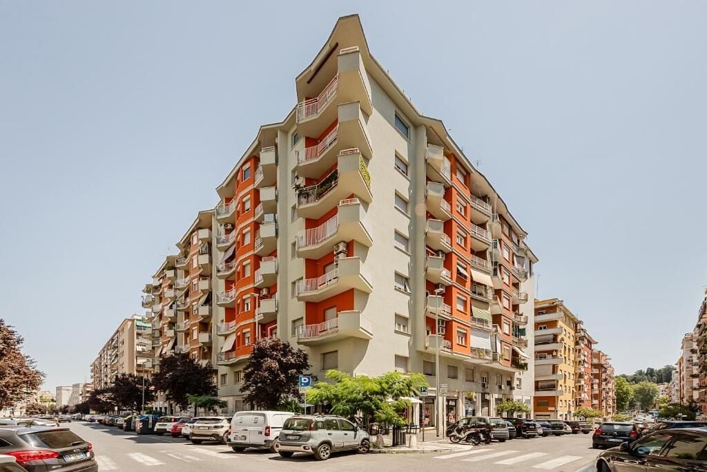 Edifici residencial de diversos pisos amb balcons, una zona d'aparcament al davant i cel blau.