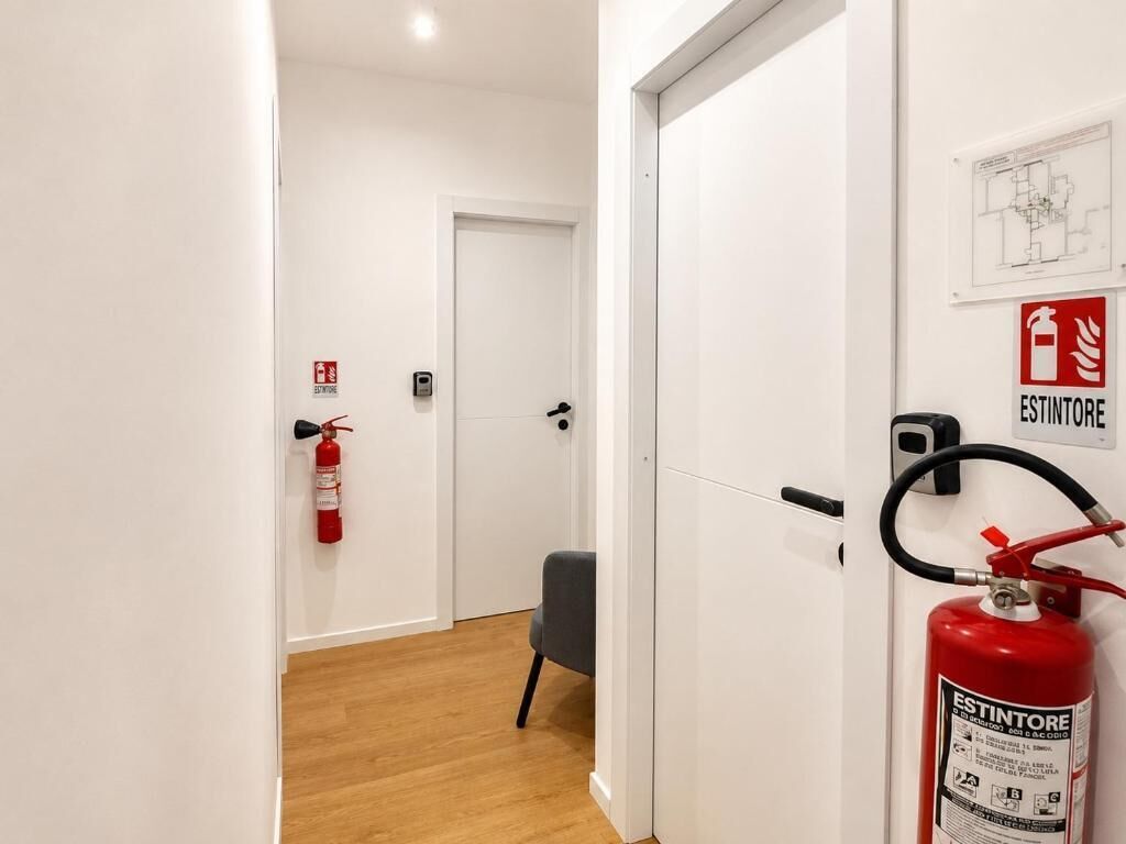A hallway with a fire extinguisher mounted on the wall, a door at the end, and a chair in the middle.