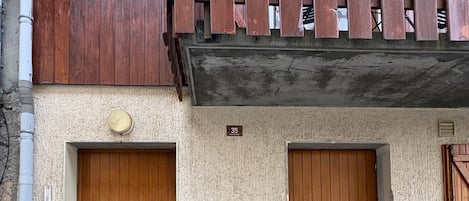 Un edificio con dos puertas de madera, un pequeño cuadro de electricidad y un balcón con barandilla de madera.