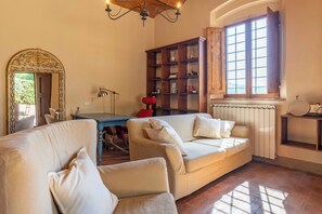 A cozy living room with a sofa, armchairs, a wooden bookshelf, and a large window. - Podere Gora (Toscana)