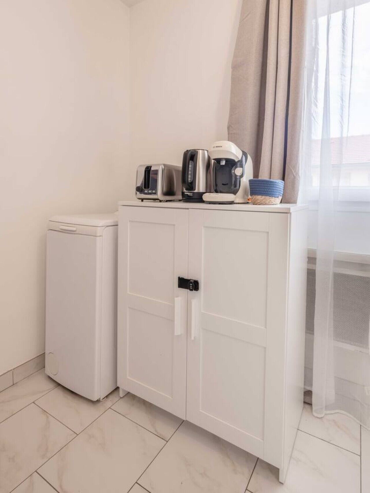 A kitchenette with a refrigerator, a toaster, a kettle, and a coffee maker on a white cabinet.