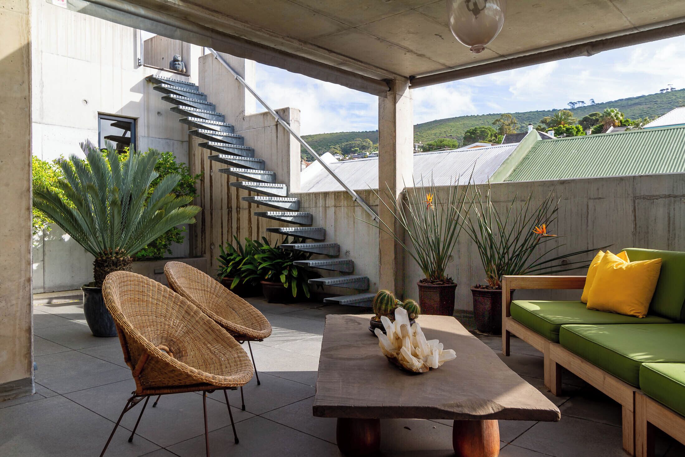 A covered outdoor patio with a wooden table, wicker chairs, a green sofa with yellow cushions, potted plants, and a staircase leading to a building.