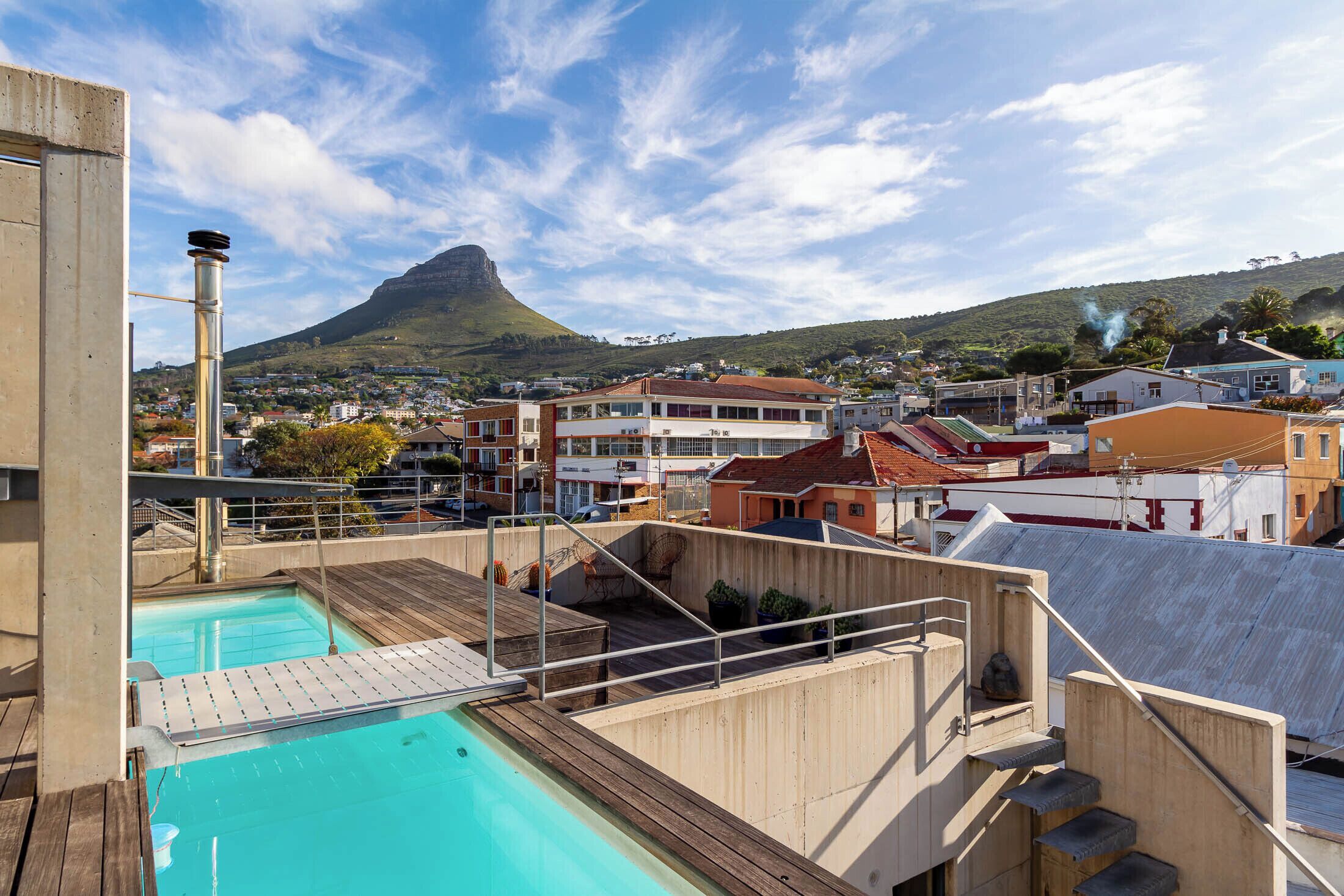 A swimming pool with a wooden deck and a view of a town with buildings and a mountain in the background.