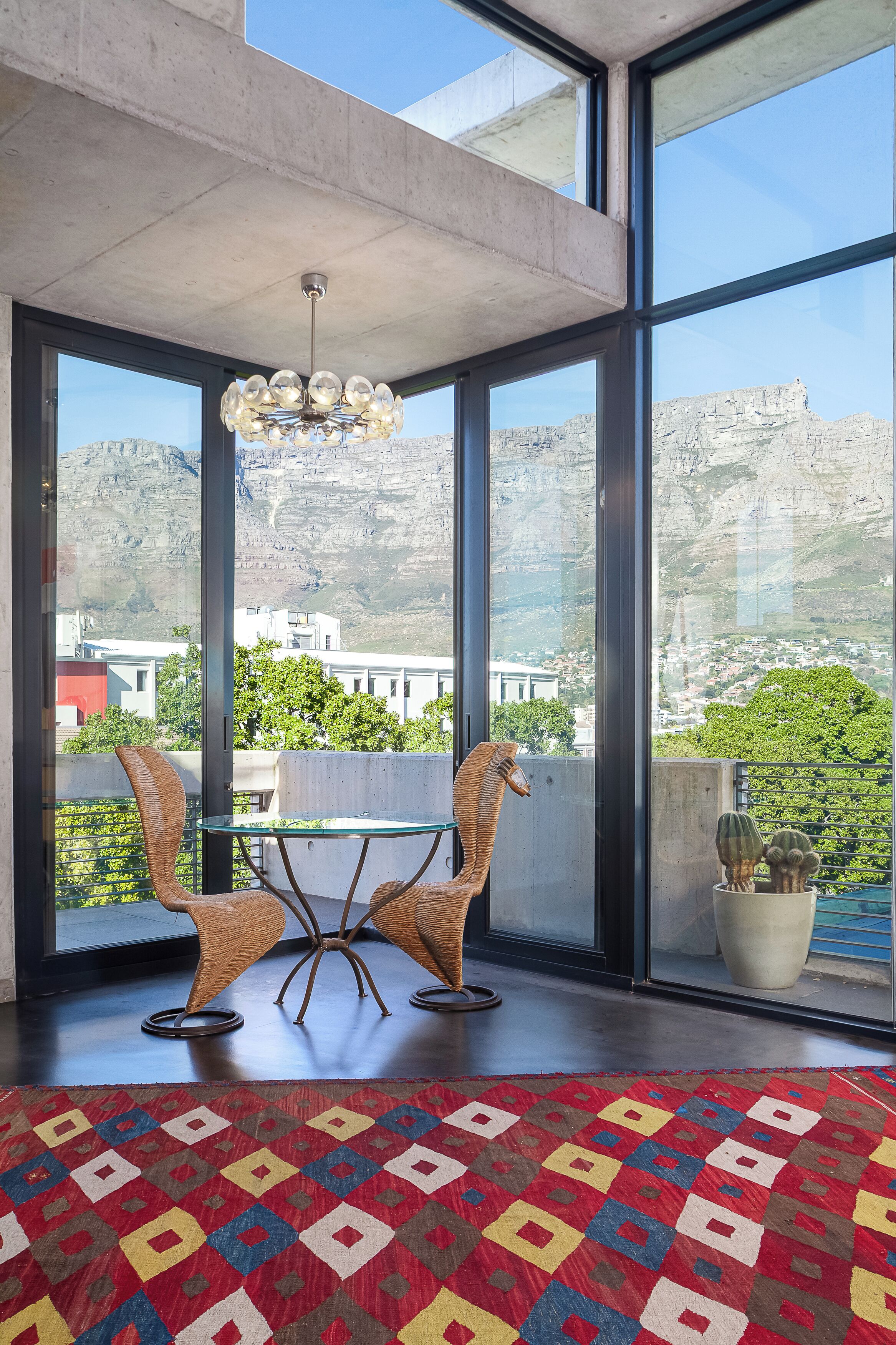 A modern interior with a glass wall, two wicker chairs, a glass table, a patterned rug, and a chandelier.