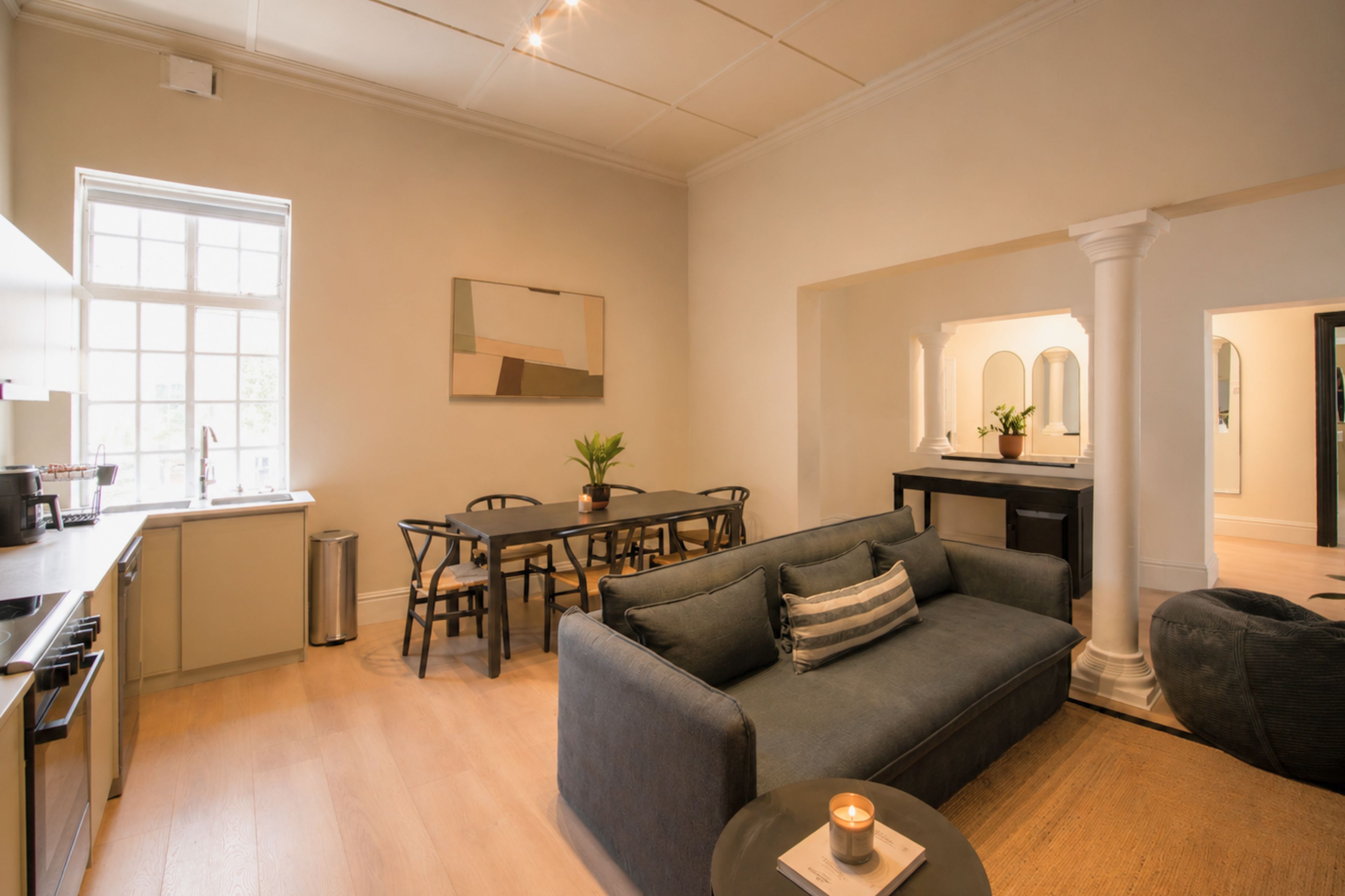 Een moderne woonkamer met een grijze bank, een eettafel met stoelen, een keukenhoek en een groot raam.