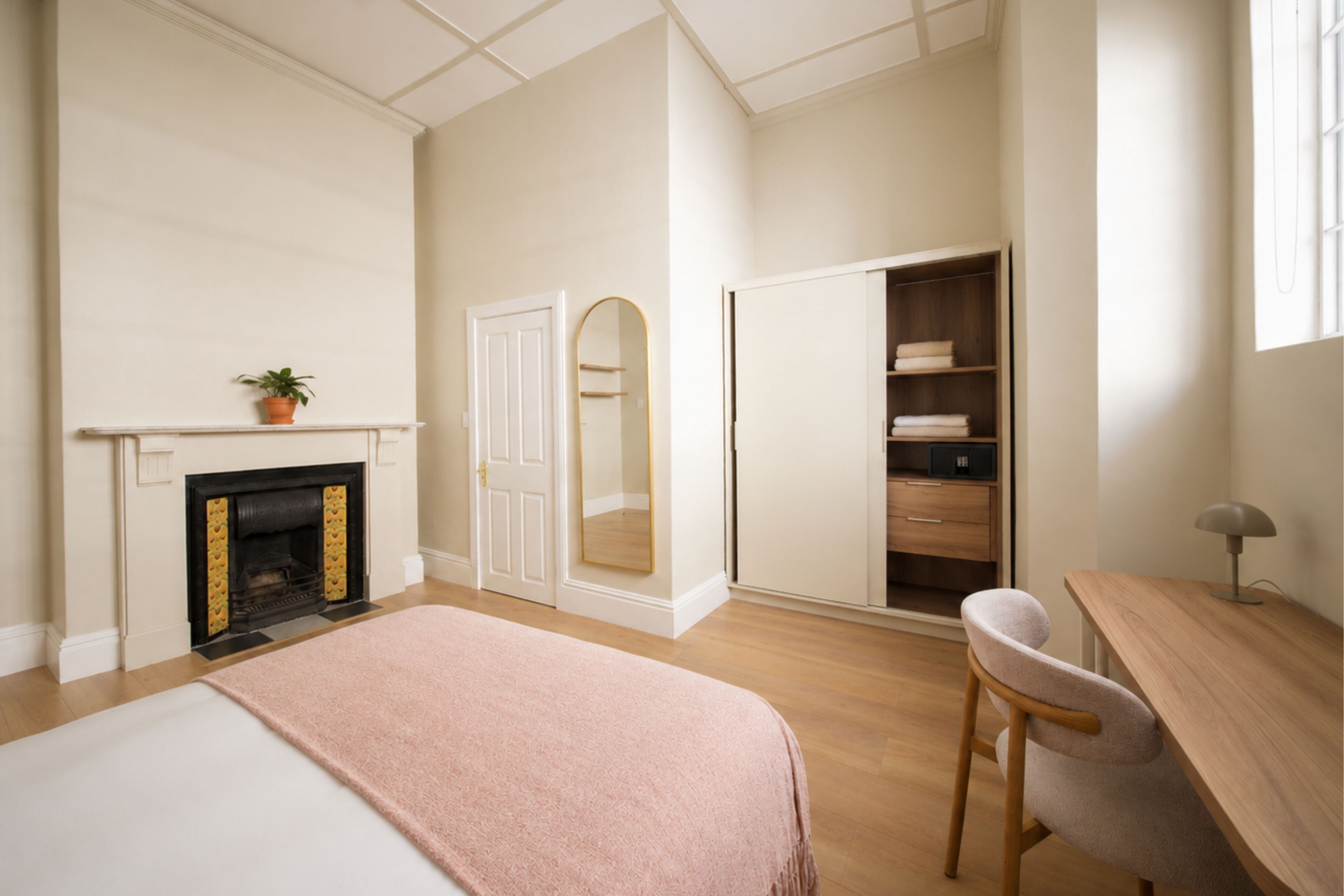 A bedroom with a bed, a desk, a chair, a wardrobe, a mirror, a fireplace, and a potted plant.