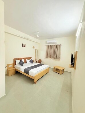 A bedroom with a bed, bedside tables, a bench, an air conditioning unit, and a window with curtains. - Hotel Krishna Garden (Valsad)