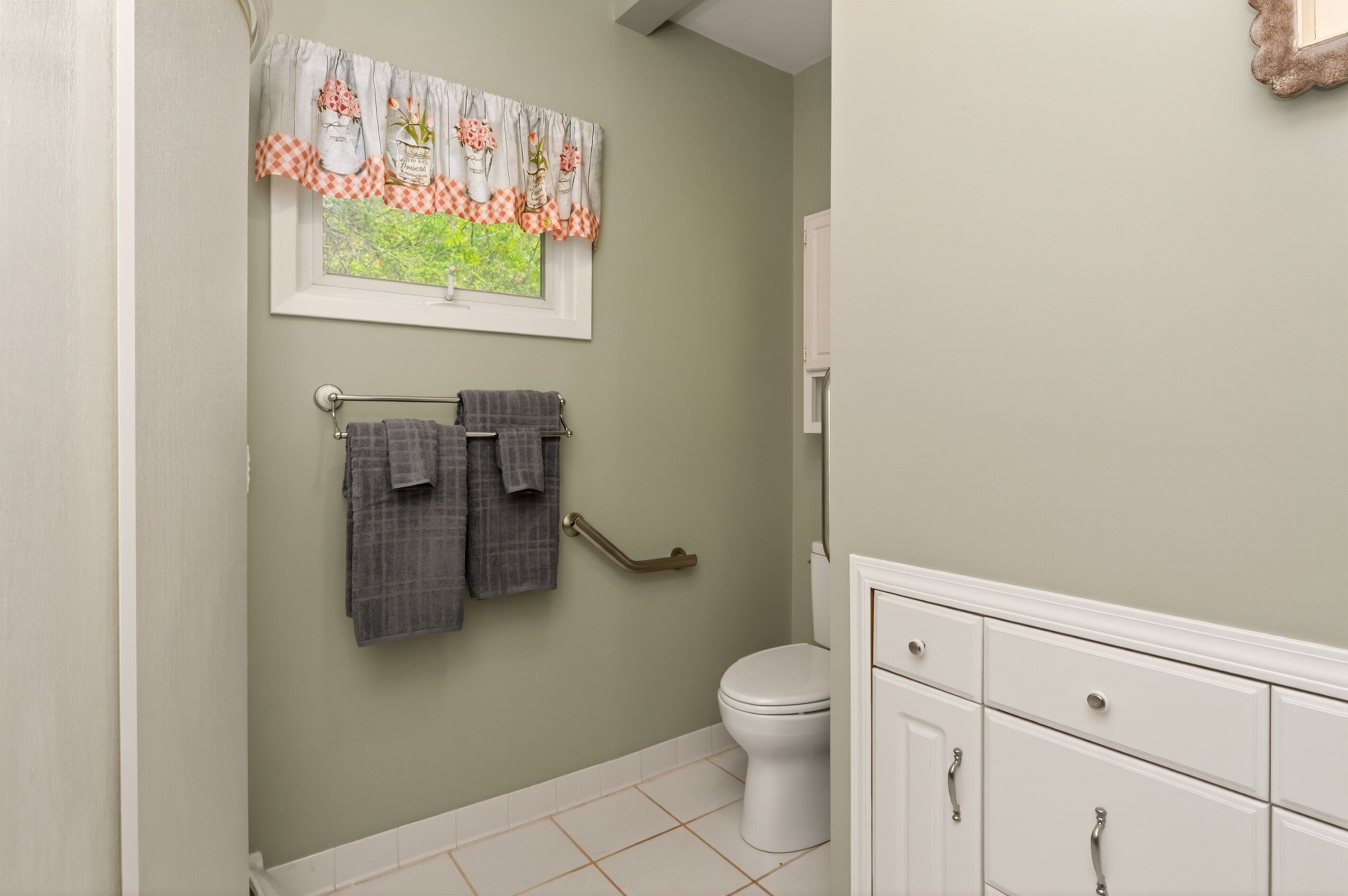 A bathroom with a toilet, a towel rack with two towels, a window with a curtain, and a cabinet with drawers.