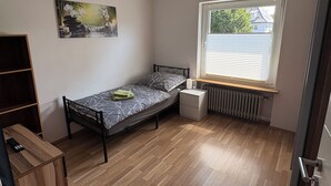 Et lille soveværelse med en enkelt seng, en radiator, et vindue med rullegardiner, et billede på væggen og trægulv.