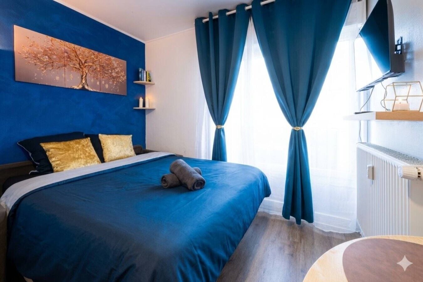 A hotel room with a bed, a painting on the wall, a window with curtains, and a bedside table.