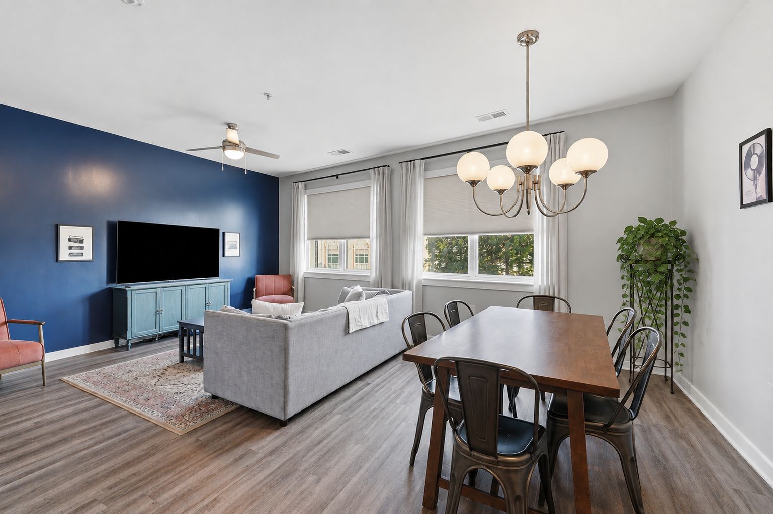 A modern living room with a flat-screen TV, a sofa, a dining table with chairs, and a ceiling fan.