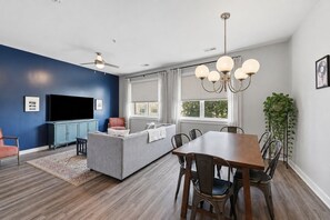 A modern living room with a flat-screen TV, a sofa, a dining table with chairs, and a ceiling fan.