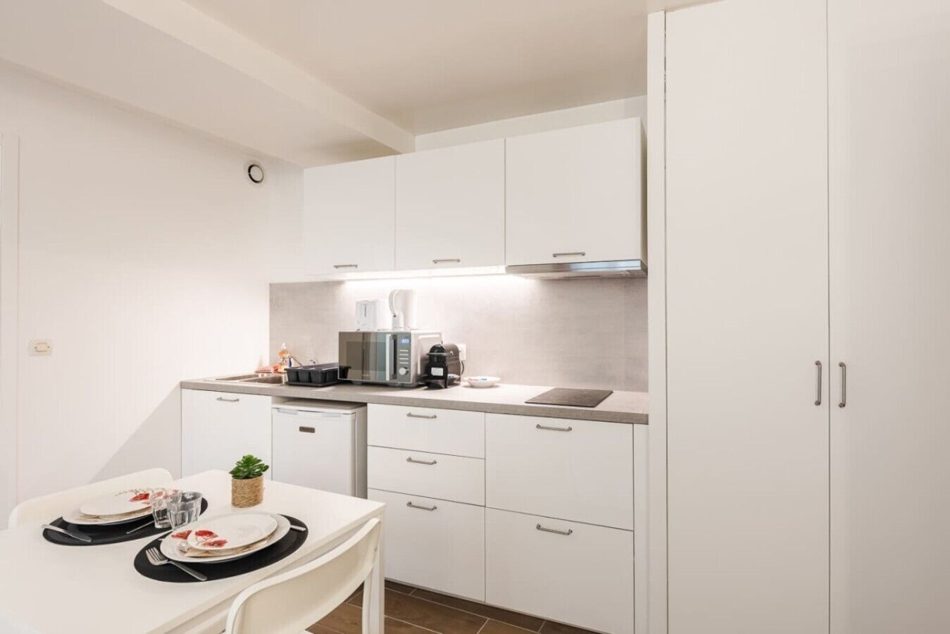 A compact kitchen with white cabinets, a dining table set for two, and a coffee machine.