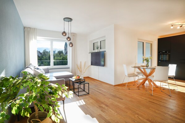 Uma sala de estar moderna com um sofá, uma mesa de jantar com cadeiras, uma TV na parede e uma janela grande com vista para árvores e edifícios.