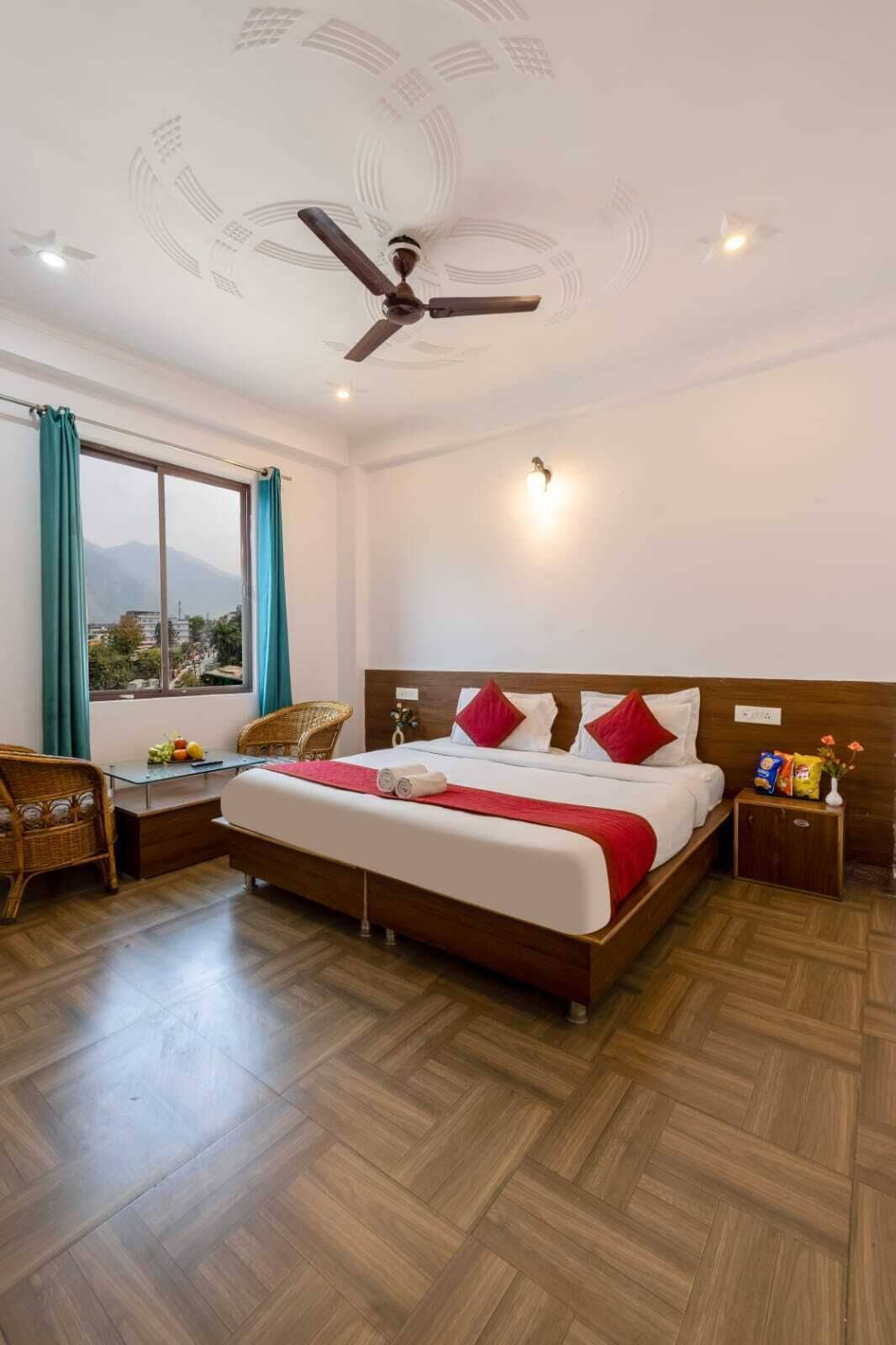 Quarto de hotel com cama, ventilador de teto, mesa de cabeceira, cadeira, mesa e janela com cortinas.