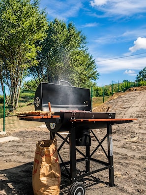 A portable barbecue grill with a metal frame and wheels, set up on a dirt ground with a bag of charcoal nearby.