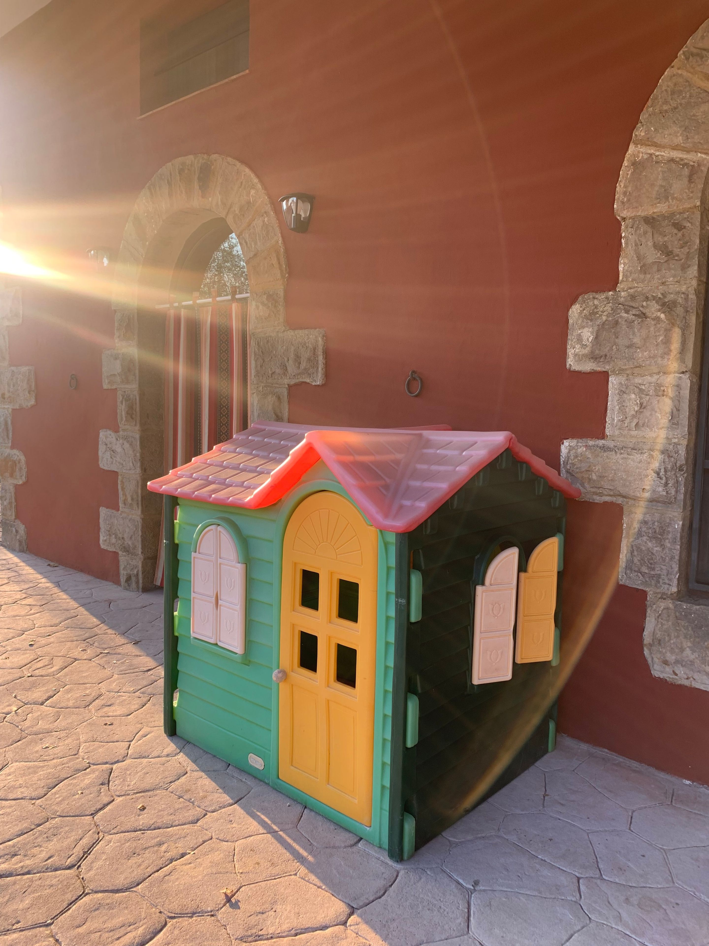Una casita de juegos pequeña y colorida con techo rojo y paredes verdes, ubicada en una zona pavimentada con muros y arcos de piedra al fondo.