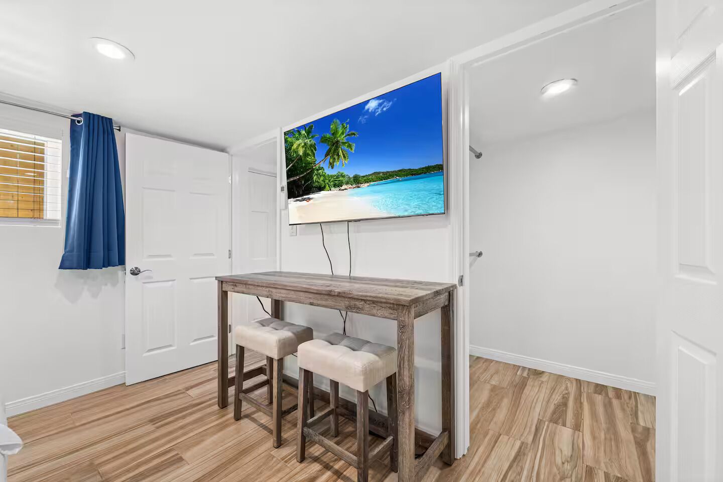 A room with a wooden table and two stools, a television mounted on the wall, and a window with blinds.