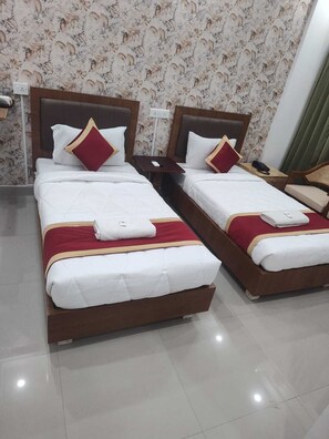 A hotel room with two beds, each with white bedding and red accents, a small bedside table with a phone, and a wall with a floral pattern. - PPH Living Sri Sai Comforts (Bengaluru)