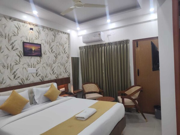 A hotel room with a bed, two chairs, a desk, a ceiling fan, and a window with curtains. - PPH Living Sri Sai Comforts (Bengaluru)