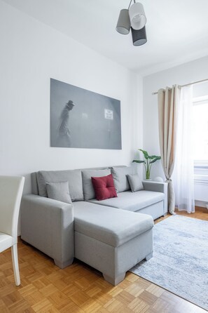 Ein modernes Wohnzimmer mit einer grauen Eckcouch, einem roten Zierkissen, einem weißen Sessel, einer grauen Pendelleuchte und einem großen abstrakten Gemälde an der Wand.