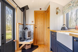 A compact kitchen with a wood-burning stove, a window with a view of trees, and a door leading to another room.