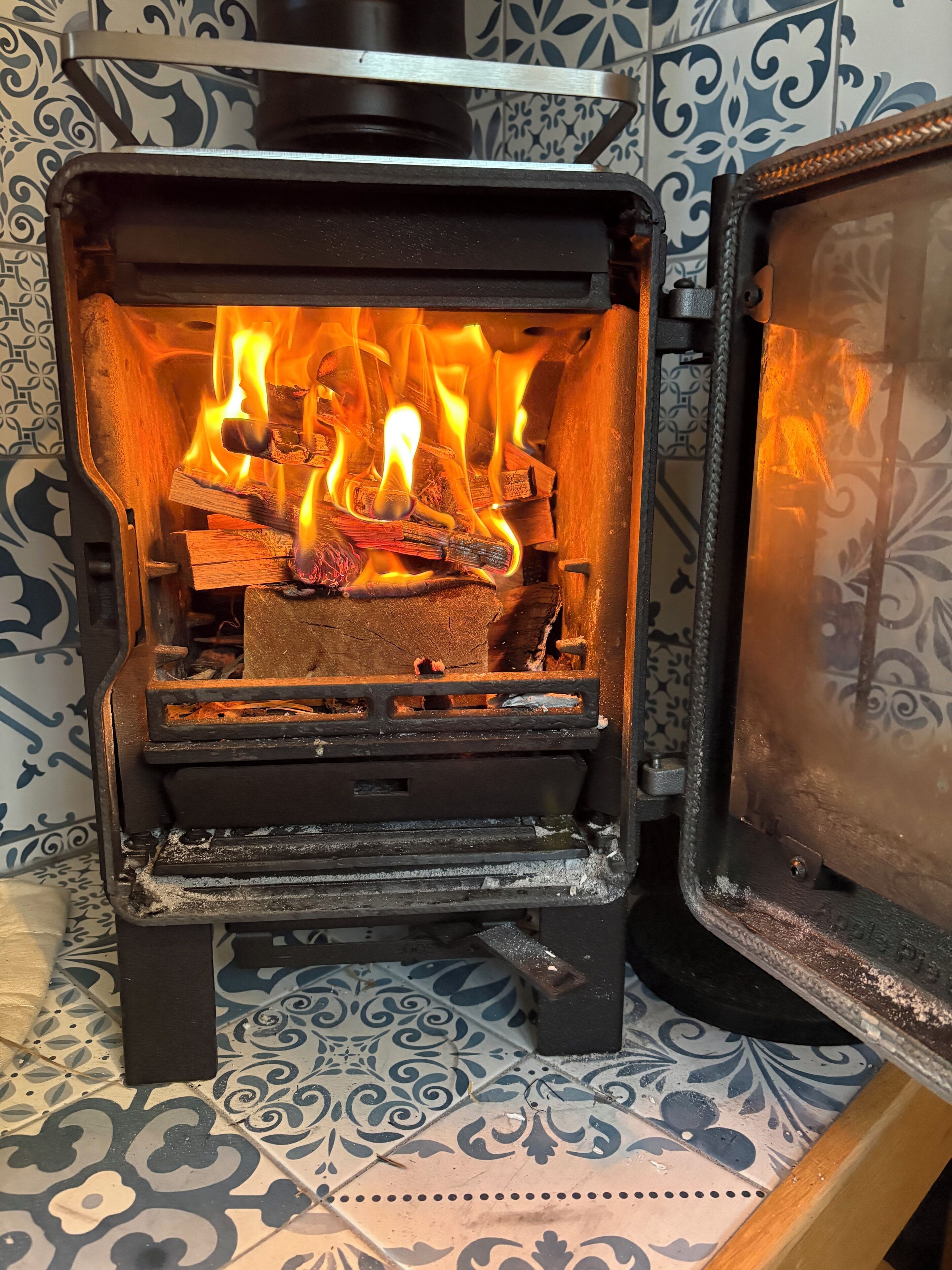 Ein Kaminofen mit sichtbaren Flammen vor einer gefliesten Wand mit dekorativem Muster.