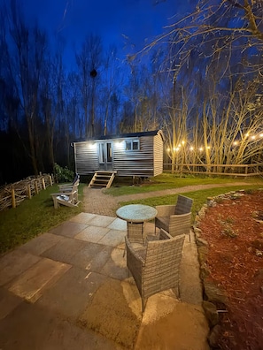 A small, single-story RV with a sliding door, a patio with outdoor furniture, and a wooden deck.
