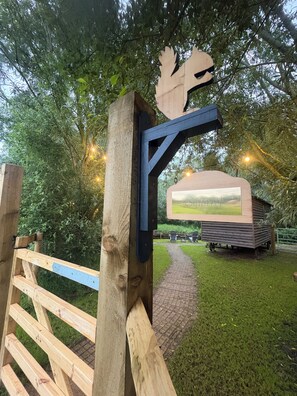 A wooden gate with a leaf-shaped sign, a pathway leading to a building with a picture of a field, and a grassy area with trees.
