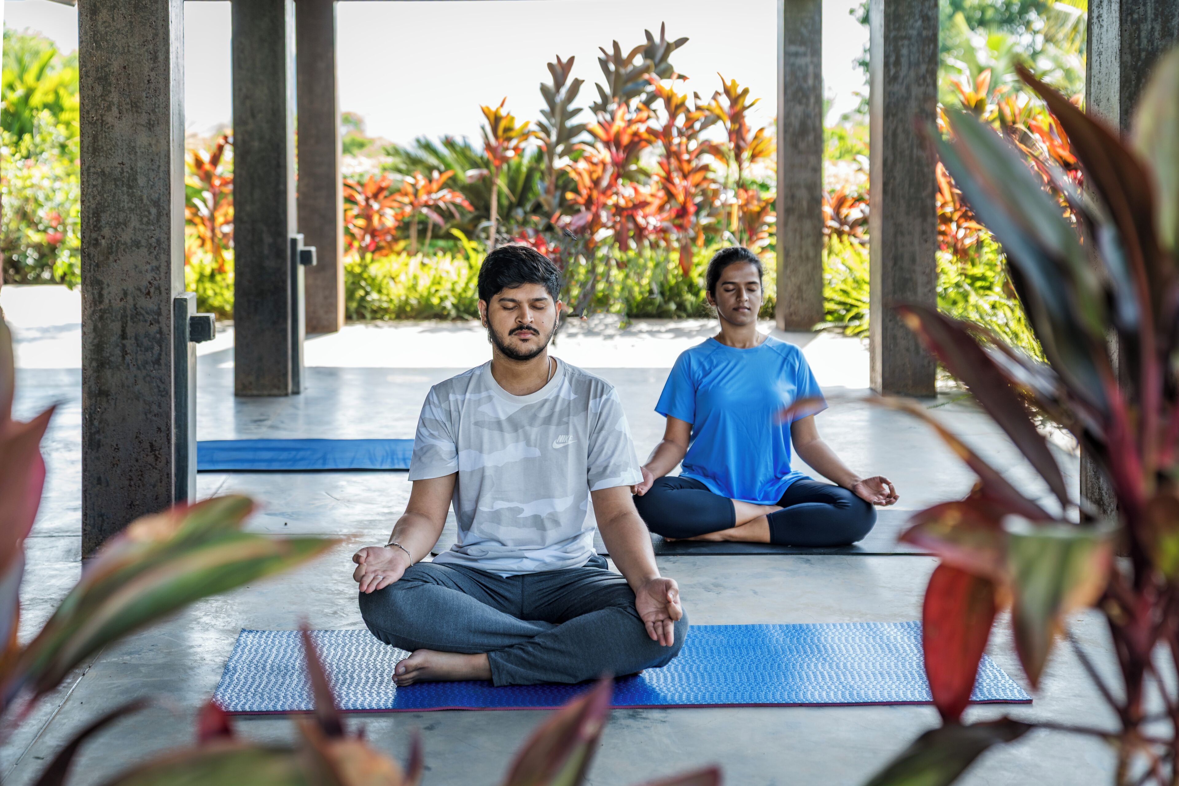 Dos personas sentadas sobre esterillas de yoga en un espacio exterior con plantas y una estructura al fondo.