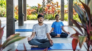 Dos personas sentadas sobre esterillas de yoga en un espacio exterior con plantas y una estructura al fondo.